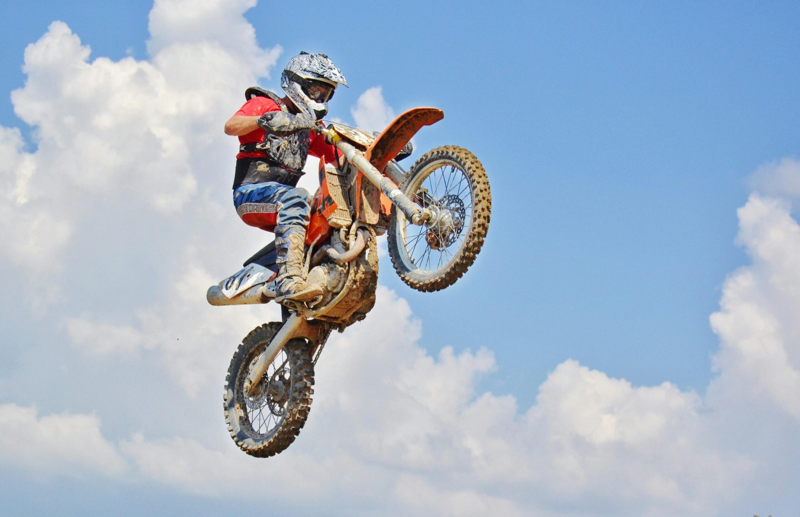 Motocross rider performs an airborne jump under a bright blue sky with clouds.